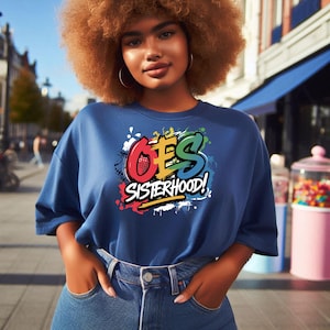 Puede incluir: Una mujer con una camiseta azul con un gráfico colorido que dice "OES Sisterhood!" El gráfico es de estilo graffiti con salpicaduras de pintura.