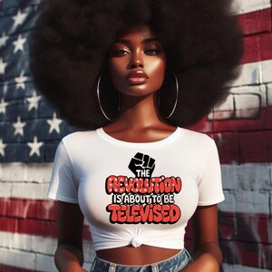 May include: A woman with an afro hairstyle is wearing a white t-shirt with a red graphic that says "The Revolution is About to Be Televised". The graphic also features a black fist.