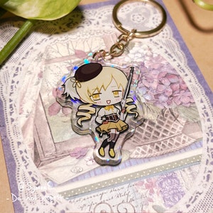 May include: A clear acrylic keychain featuring a cartoon character with blonde hair, a black beret, and a yellow dress. The character is holding a rifle and is smiling. The keychain has a holographic effect.