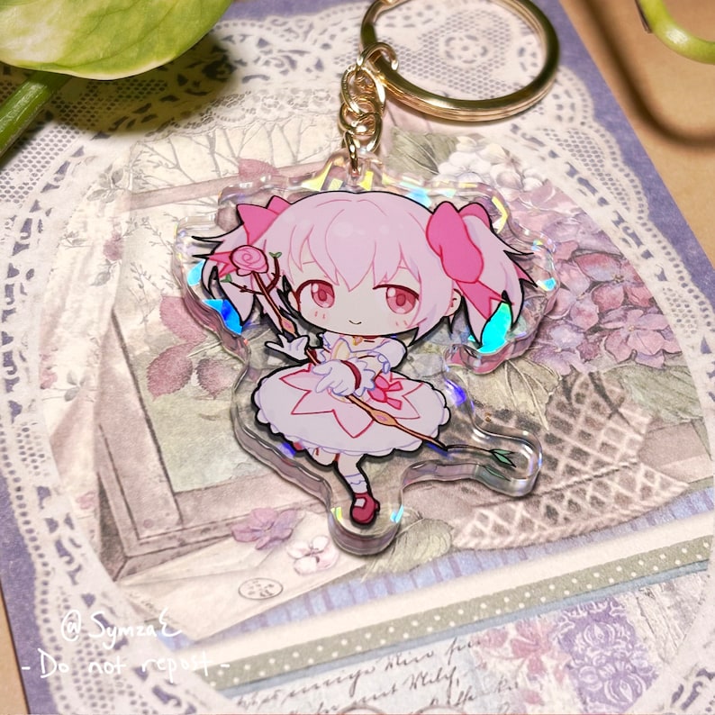 May include: A pink and white anime character keychain with a pink bow and a rose staff. The keychain is made of clear acrylic with a holographic finish.