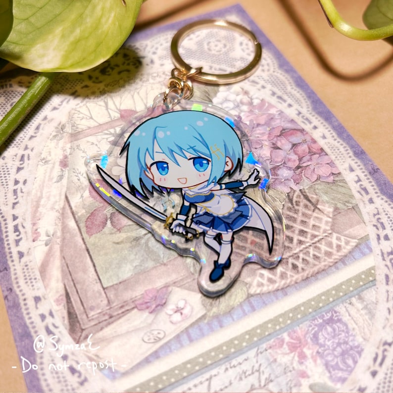 May include: A blue and white anime character keychain with a sword. The character is a young girl with blue hair and a blue and white dress. The keychain is made of clear acrylic with a holographic finish.