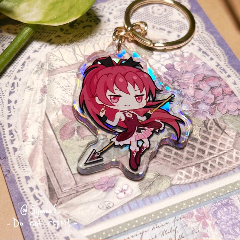 May include: A holographic keychain featuring a cartoon character with red hair, a red dress, and a spear. The character is holding a spear and has a serious expression on her face. The keychain is made of clear acrylic and has a gold metal ring.