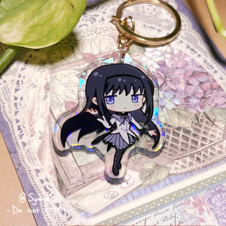 May include: A holographic keychain featuring a cartoon anime character with black hair, a white and purple dress, and black boots. The character is holding a wand in her right hand and has a surprised expression on her face.