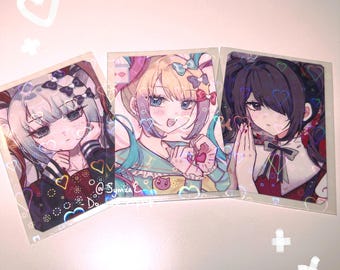 May include: Three rectangular anime-style art prints featuring illustrated characters with colorful hair and detailed outfits. The prints are encased in clear protective sleeves and feature a holographic overlay with heart and star patterns.