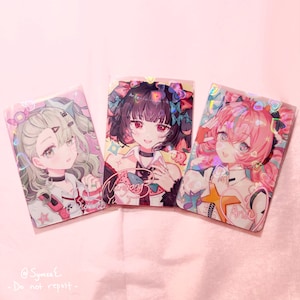 May include: Three anime-style character cards with holographic accents. Each card features a different girl with unique hairstyles and outfits. The cards are set against a soft pink background. The cards have the names "Yumea", "Nangong", and "Aria" on them.