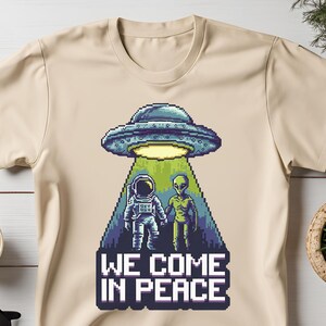 May include: A beige t-shirt with a pixelated graphic of a UFO, an astronaut, and an alien. The text "WE COME IN PEACE" is printed below the graphic.