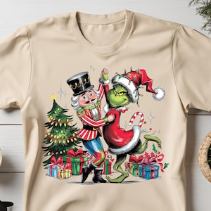 May include: A beige t-shirt featuring a festive Christmas design with the Grinch and the Nutcracker dancing together. The Grinch is wearing a red Santa hat and a green suit, while the Nutcracker is wearing a traditional red and gold costume. There is a Christmas tree and presents in the background.