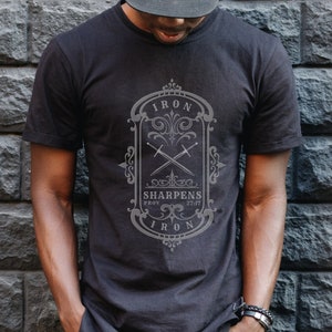 Peut inclure: Un t-shirt noir avec un motif graphique gris représentant une épée stylisée et le texte "Iron Sharpens Iron" dans une police d'écriture d'inspiration vintage.