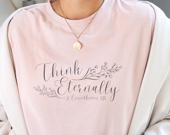 以下が含まれることがあります： 花柄のデザインで「Think Eternally」というテキストと聖書の引用「コリントの信徒への手紙第二 4:18」が書かれたピンクのTシャツです。