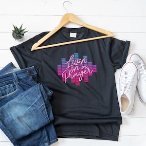 Könnte beinhalten: Schwarzes T-Shirt mit einer pinkfarbenen und blauen Grafik, auf der "Livin' on a Prayer" steht. Das Shirt hängt an einem Holzbügel und liegt auf einer weißen Holzoberfläche.