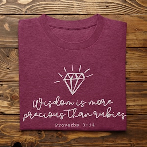 Puede incluir: Una camiseta color granate con un gráfico de diamante blanco y el texto "Wisdom is more precious than rubies" y "Proverbs 3:14".
