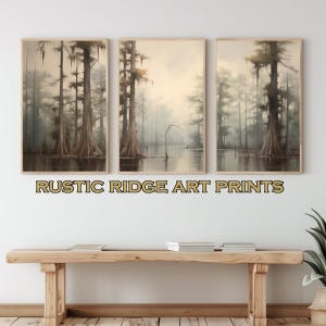 May include: Three framed landscape paintings of a misty swamp with tall trees. The paintings are hung on a white wall above a wooden bench. The text "RUSTIC RIDGE ART PRINTS" is displayed below the paintings.