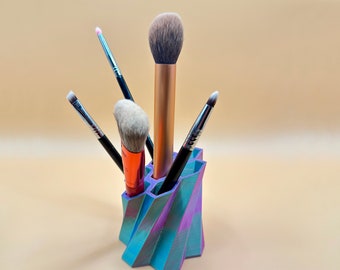 May include: A multi-colored, 3D-printed makeup brush holder with a geometric design. The holder contains several makeup brushes with various handle colors and brush head shapes. The holder's colors include blue, purple, and pink.