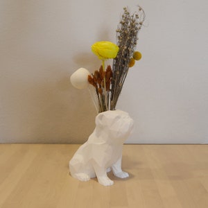May include: A white, geometric bulldog-shaped vase holding dried flowers. The arrangement includes a large yellow flower, white spherical accents, and various dried stems. The vase sits on a light wood surface.