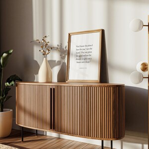 May include: A wooden cabinet with a curved front and vertical slats. The cabinet has two doors and sits on black metal legs. A framed print with a quote about plans and hope is on the wall behind the cabinet. A vase with dried branches is on the cabinet. A floor lamp with white globes and gold accents is in the background.
