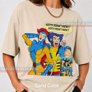 May include: A sand-colored t-shirt featuring a graphic print of two comic book characters. One character has red hair and a blue and yellow suit, while the other has blue hair and a yellow and blue suit, holding a microphone with the text SCOTTY DOESN'T KNOW!!!