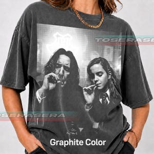 May include: A charcoal gray short-sleeve t-shirt featuring a black and white graphic print of two individuals smoking. The shirt has a crew neckline and a relaxed fit. The text "Graphite Color" is printed at the bottom of the shirt.