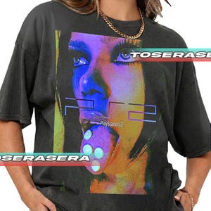 Puede incluir: Camiseta gris oscuro con un retrato estilizado y vibrante en tonos azules, morados y amarillos. El diseño incluye el texto "PS2" y "PlayStation 2". La camiseta tiene un corte holgado y mangas cortas.