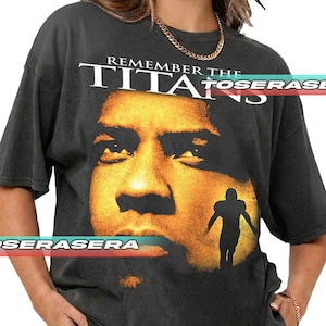 Könnte beinhalten: Dunkelgraues T-Shirt mit dem Schriftzug "REMEMBER THE TITANS" über einem großen Porträt eines Mannes. Eine Silhouette eines Footballspielers befindet sich darunter. Das T-Shirt hat kurze Ärmel.