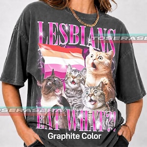 May include: A charcoal grey short-sleeve t-shirt featuring a graphic print of three cats with surprised expressions. Above the cats is the text LESBIANS and a lesbian pride flag. Below the cats is the text EAT WHAT?! and the words Graphite Color.