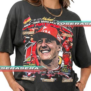 Könnte beinhalten: Dunkelgraues T-Shirt mit einer Grafik von Michael Schumacher mit roter Kappe, der Nummer 1 und den Jahren 2000, 2001, 2003, 2004. Das T-Shirt hat einen Vintage-Look.