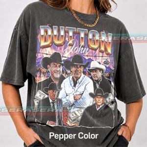 Könnte beinhalten: Ein anthrazitfarbenes Kurzarm-T-Shirt mit einem Grafikdruck von John Dutton aus Yellowstone. Das Design enthält den Text DUTTON John, mehrere Bilder eines Mannes mit Cowboyhut, einen Cowboystiefel, Geldbündel und einen Hubschrauber.