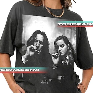 以下が含まれることがあります： 2人の喫煙者の白黒グラフィックが特徴の濃い灰色のTシャツ。「TOSERASE」の文字が画像の上に、「OSERASERA」が下にあります。このシャツは半袖で、ゆったりとしたフィット感です。