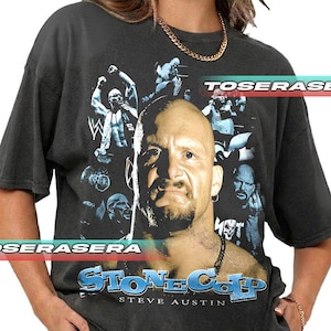 May include: Dark gray t-shirt featuring a graphic of a bald man with a goatee, surrounded by images of other men. The text "STONE COLD" and "STEVE AUSTIN" are printed in blue and white. The shirt also has the text "TOSERASE" and "SERASERA".