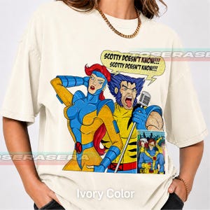 Op de afbeelding: Een ivoorkleurig t-shirt met een grafische print van twee stripfiguren, één met rood haar en één met blauw haar, die in een microfoon zingen. Een tekstballon boven hen bevat de tekst "SCOTTY DOESN'T KNOW!!! SCOTTY DOESN'T KNOW!!!".