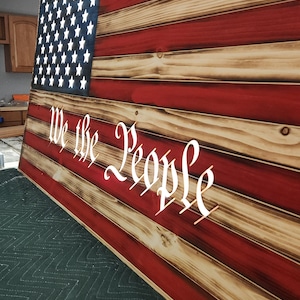 May include: A wooden American flag with red, white, and blue stripes and a field of blue stars. The words "We the People" are inscribed in white on the flag.