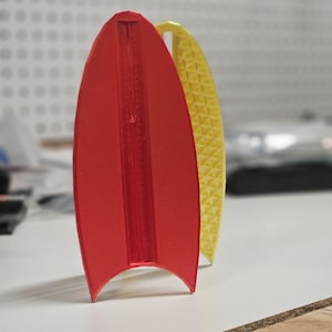 May include: Two 3D printed surfboard fin shapes, one red and one yellow. The red fin has a smooth surface and the yellow fin has a textured surface.