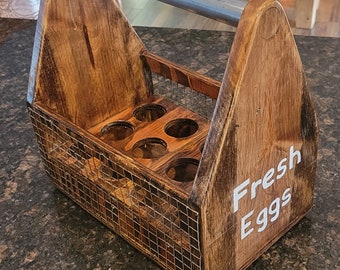 Puede incluir: Porta huevos de madera con asa metálica y laterales de malla metálica. El soporte está teñido de un marrón cálido y tiene las palabras "Fresh Eggs" en letras blancas. Tiene seis compartimentos circulares.