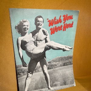 May include: A vintage postcard featuring a black and white photo of a couple on a beach. The man is holding the woman in his arms. The text on the postcard reads "Wish You Were Here."