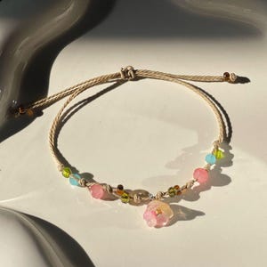 May include: A delicate bracelet featuring a beige cord with colorful beads and a flower charm. The bracelet includes pink, green, and blue beads, and a translucent pink flower pendant. The bracelet is displayed on a white, decorative plate.