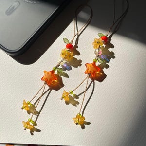 Puede incluir: Dos colgantes decorativos con cuentas en forma de estrella naranja, cuentas en forma de flor amarilla, pequeñas cuentas en forma de hoja verde, cuentas transparentes facetadas y cuentas redondas rojas, ensartadas en cordones beige.