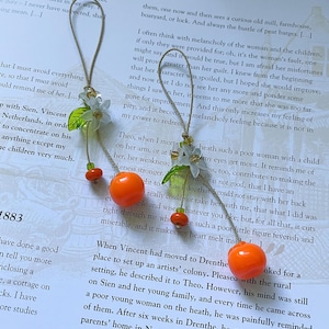 May include: Two hanging ornaments featuring a bright orange spherical bead, a smaller orange bead, a translucent white flower, and a green leaf, all suspended from a thin tan string. The ornaments are arranged vertically with decorative clear beads.