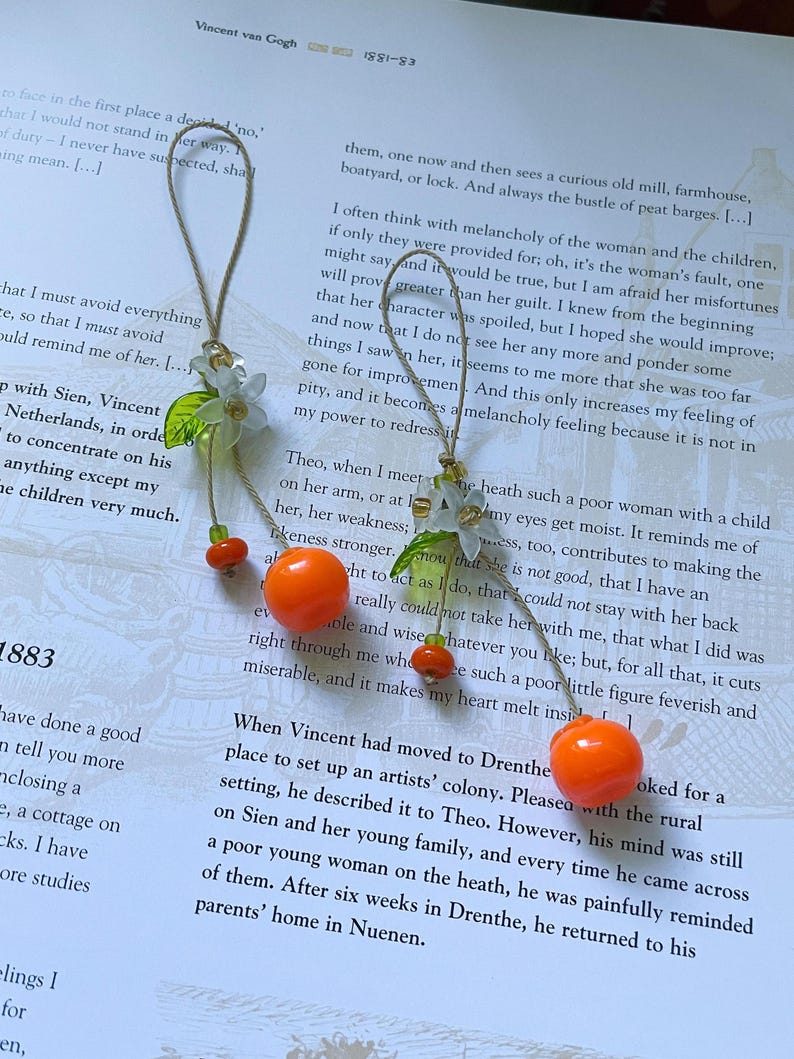 May include: Two hanging ornaments featuring a bright orange spherical bead, a smaller orange bead, a translucent white flower, and a green leaf, all suspended from a thin tan string. The ornaments are arranged vertically with decorative clear beads.