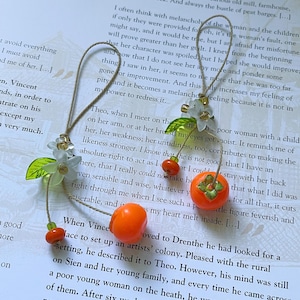 Handmade phone charm/keychain featuring two miniature bright orange persimmons crafted from clay, paired with translucent white flowers, green leaves, and small glass beads. The charm is attached to a beige cord, creating a soft, cozy aesthetic inspired by nature and fruit blossoms.