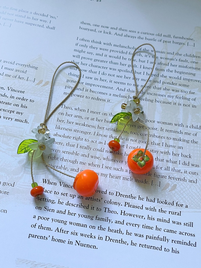 Handmade phone charm/keychain featuring two miniature bright orange persimmons crafted from clay, paired with translucent white flowers, green leaves, and small glass beads. The charm is attached to a beige cord, creating a soft, cozy aesthetic inspired by nature and fruit blossoms.