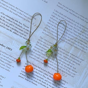 May include: Two hanging ornaments featuring a light brown string loop, a cluster of small translucent white flower petals with green leaves, and a single bright orange spherical bead dangling at the end of each piece, resting on a printed page.