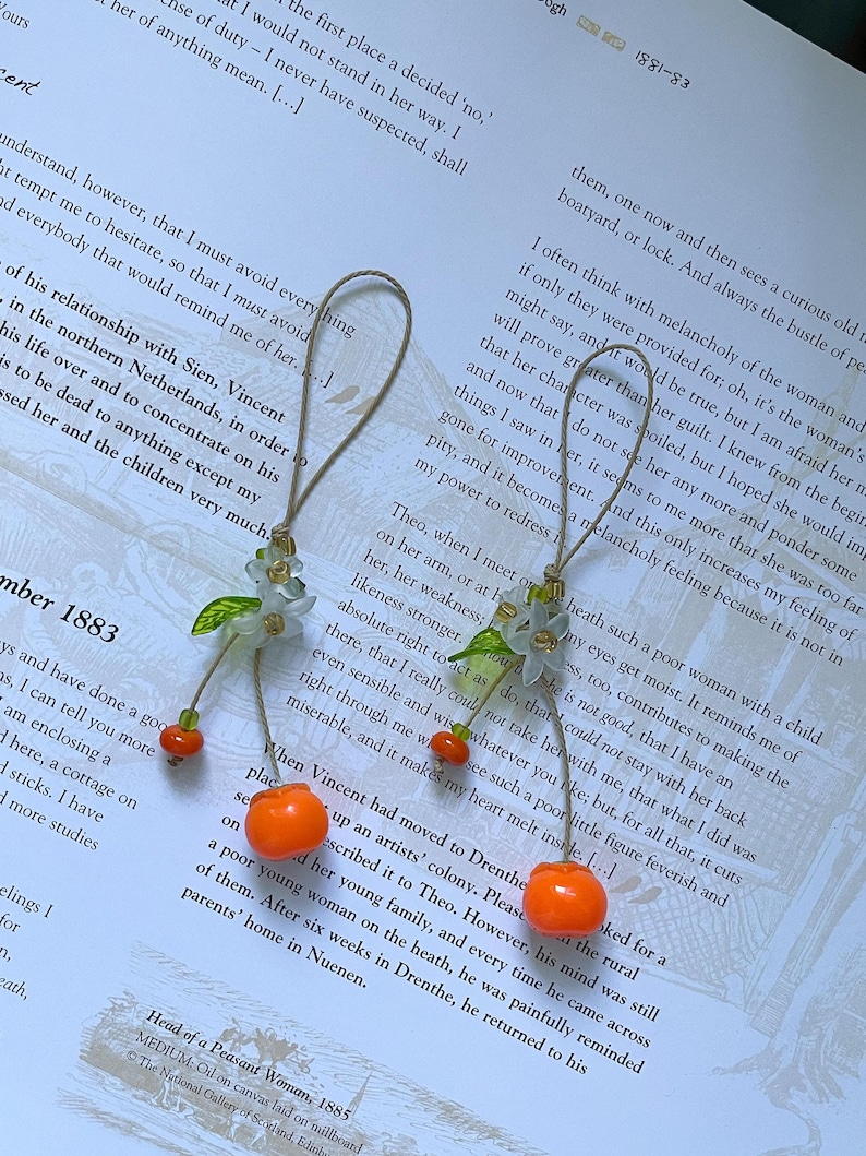 May include: Two hanging ornaments featuring a light brown string loop, a cluster of small translucent white flower petals with green leaves, and a single bright orange spherical bead dangling at the end of each piece, resting on a printed page.