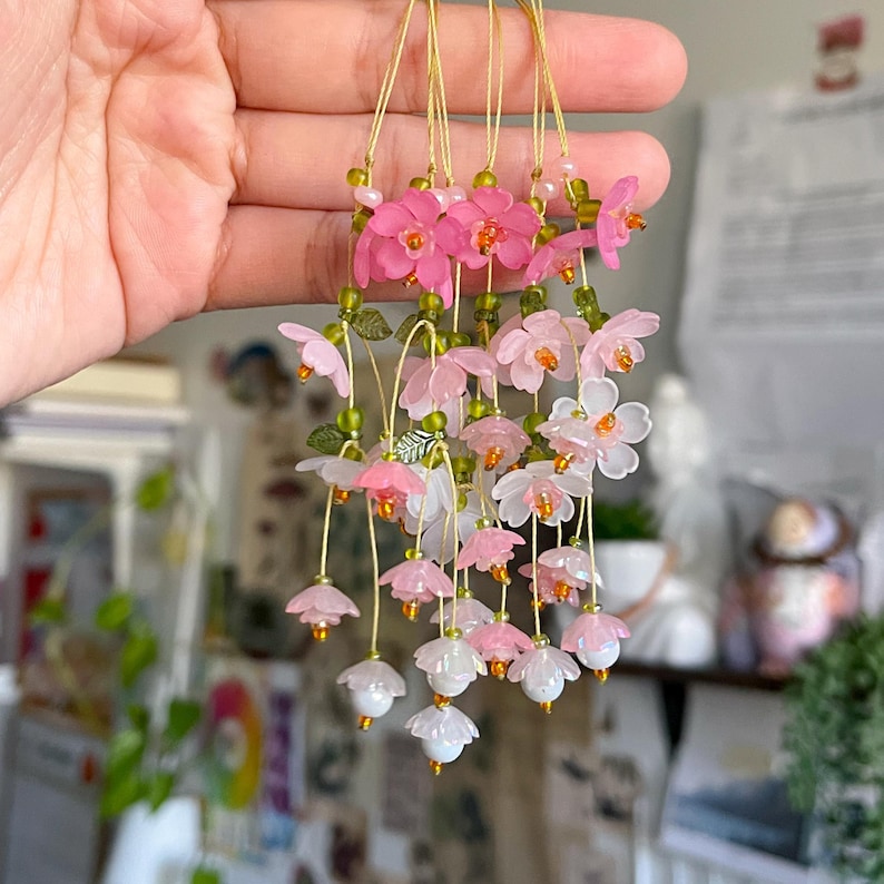 May include: Delicate floral ornaments in shades of pink and white, strung together with gold-colored wire. Each flower has a small, orange center and is accented with green beads and leaves. The ornaments are held up by a hand.