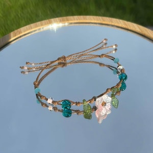 May include: A delicate bracelet made with brown cord, green and blue glass beads, and a pink flower charm.