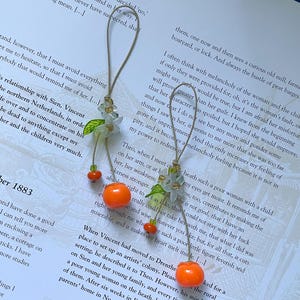 May include: Two hanging ornaments featuring light green flower-shaped beads with small green leaves, suspended by thin tan cords. Each ornament includes a bright orange spherical bead and a smaller orange bead, creating a dangling fruit-like design.