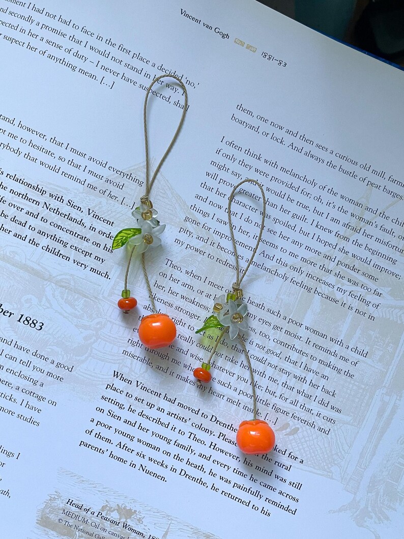 May include: Two hanging ornaments featuring light green flower-shaped beads with small green leaves, suspended by thin tan cords. Each ornament includes a bright orange spherical bead and a smaller orange bead, creating a dangling fruit-like design.