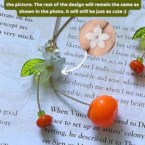 May include: A phone charm featuring a translucent white flower, a green leaf, and two orange fruit beads on a tan cord. A circular inset shows a detailed view of the white flower. Text on the image states: Please note: Due to a material shortage, the white Flower will be replaced with a different white flower, as shown in the picture. The rest of the design will remain the same as shown in the photo. It will still be just as cute :)