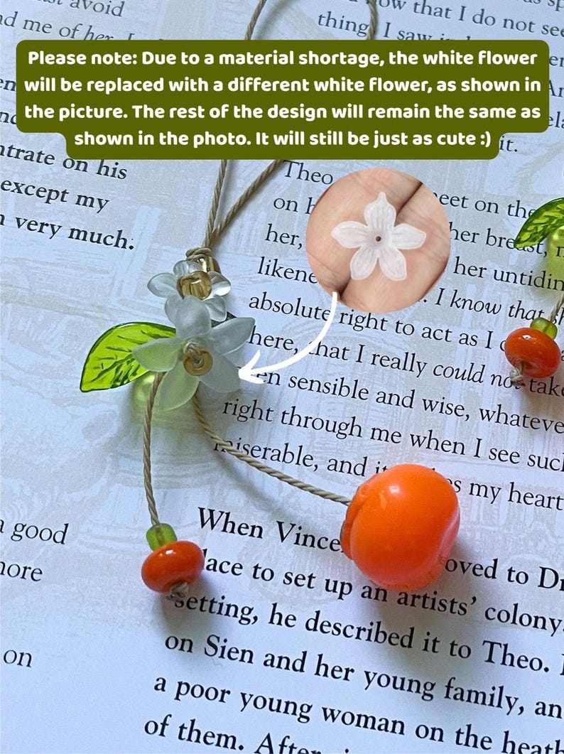May include: A phone charm featuring a translucent white flower, a green leaf, and two orange fruit beads on a tan cord. A circular inset shows a detailed view of the white flower. Text on the image states: Please note: Due to a material shortage, the white Flower will be replaced with a different white flower, as shown in the picture. The rest of the design will remain the same as shown in the photo. It will still be just as cute :)