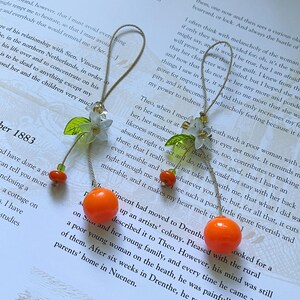 May include: Two hanging ornaments featuring a bright orange spherical bead, a small green leaf-shaped bead, and a white flower-shaped bead. Each ornament is suspended by a thin tan cord loop, accented with small translucent green and gold beads.