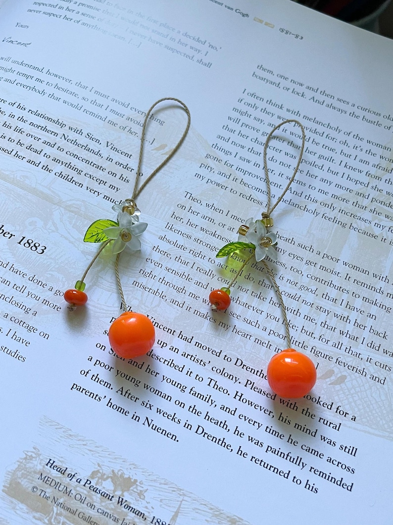 May include: Two hanging ornaments featuring a bright orange spherical bead, a small green leaf-shaped bead, and a white flower-shaped bead. Each ornament is suspended by a thin tan cord loop, accented with small translucent green and gold beads.
