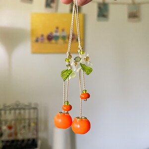 May include: A hanging ornament featuring two bright orange fruit charms suspended from thin beige cords. The design includes small translucent white flower blossoms, green glass leaves, and tiny green and orange accent beads along the strings.